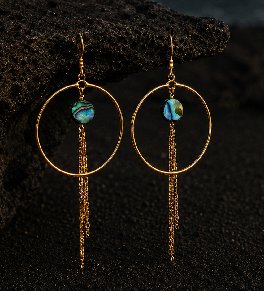A pair of gold hoop dangle earrings with small iridescent blue-green beads and long chain drops, photographed against a dark volcanic beach background with a rock and ocean waves.