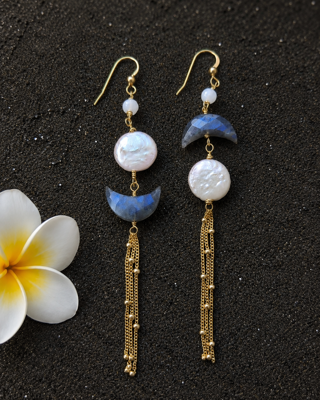Pair of earrings with gold hooks, white beads, blue crescent moons, and gold tassels on a dark background with a white flower.