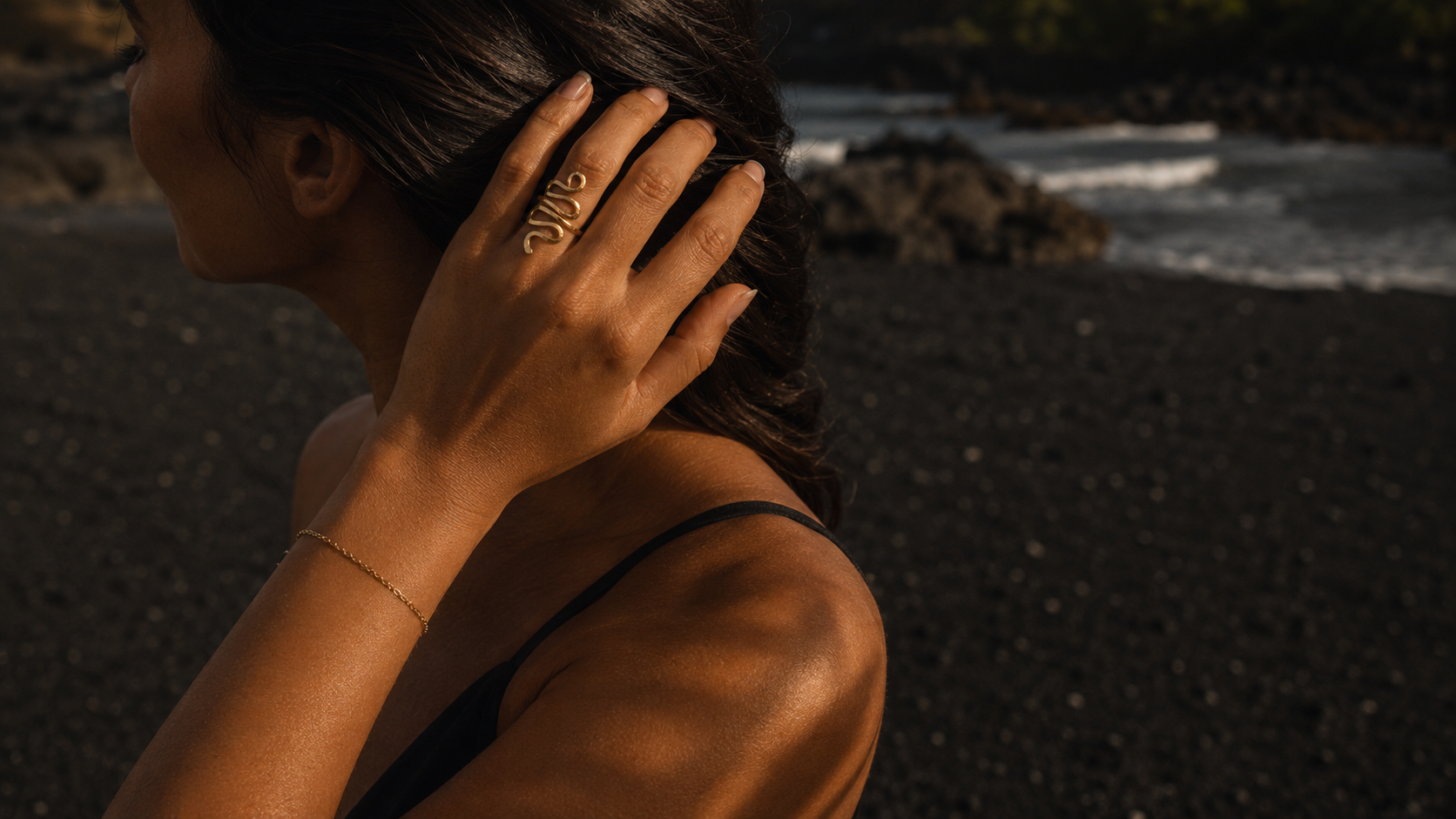 A woman on a black sand beach at sunset wearing a delicate gold chain bracelet on her wrist and a gold ring on her finger, with ocean waves and dark rocks in the background.